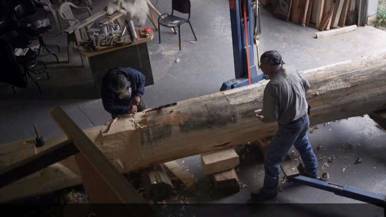 a gif from a short film about a Huna Tlingit ancestral tribal house being constructed, showing two artisans working on carving a totem pole in a workshop