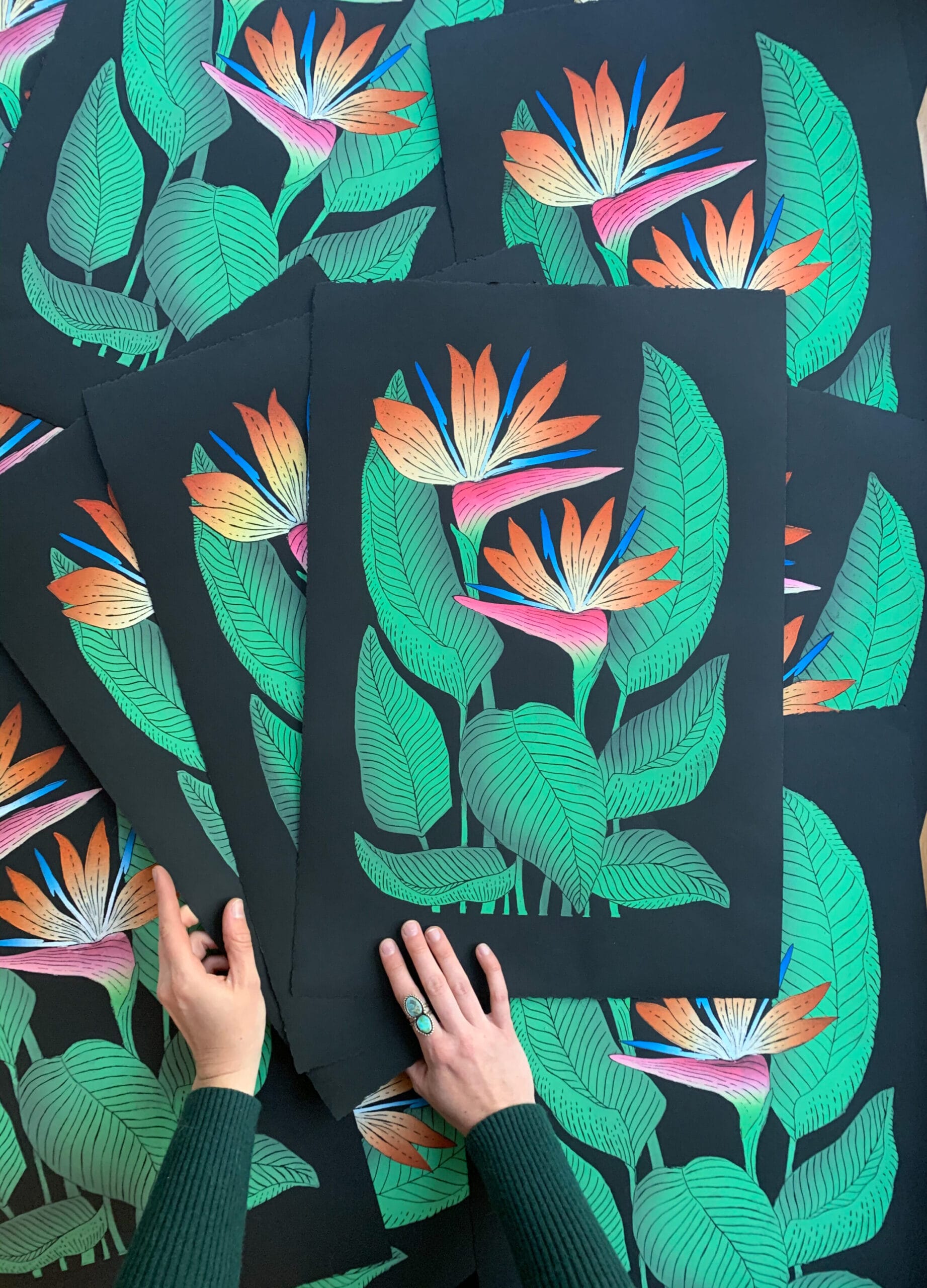 two hands touch the corners of a group of block prints of pink tropical flowers on a black background