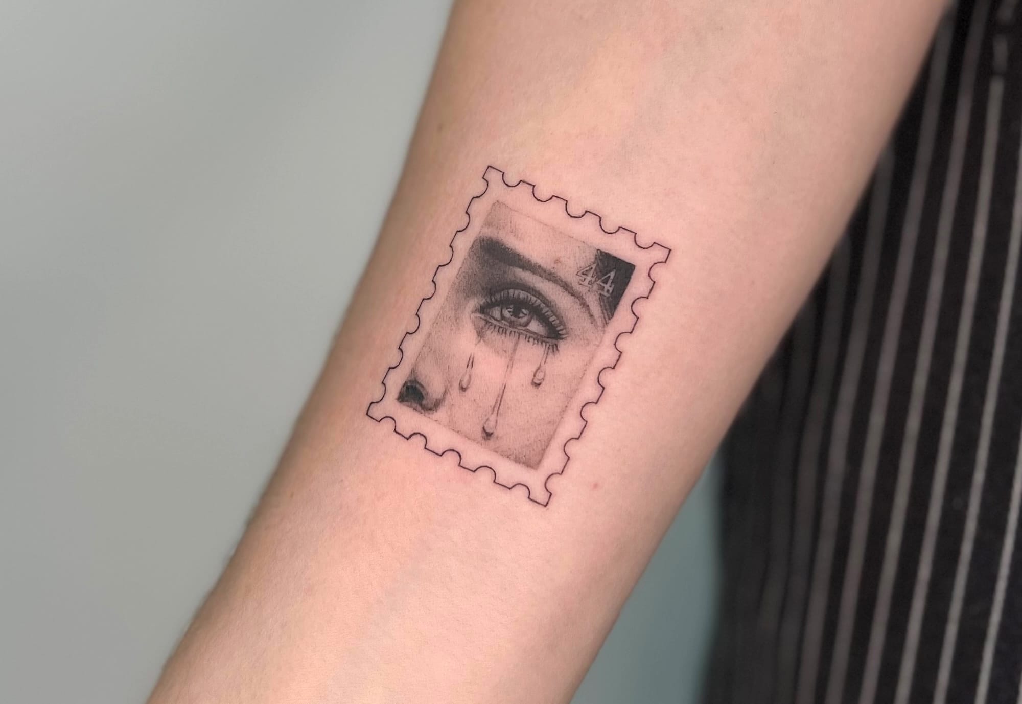 a photograph of a postage stamp-shaped tattoo on a forearm, containing a compositional detail of a woman's eye with tears rolling down her cheek