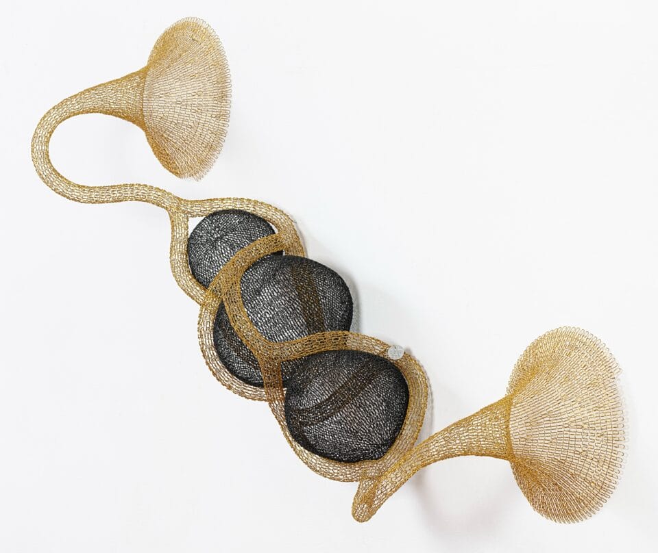 a gold and black abstract woven metal sculpture with tubes and circular forms on a wall