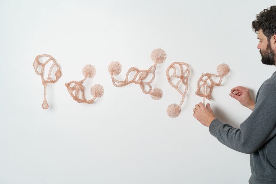 a white man touches a series of red abstract woven metal sculptures with tubes and circular forms on a wall