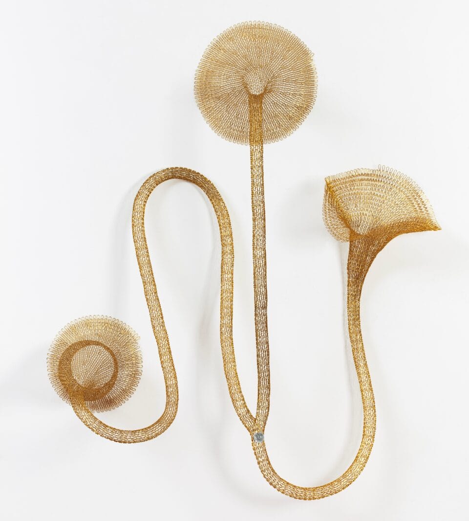 a gold abstract woven metal sculpture with tubes and circular forms on a wall