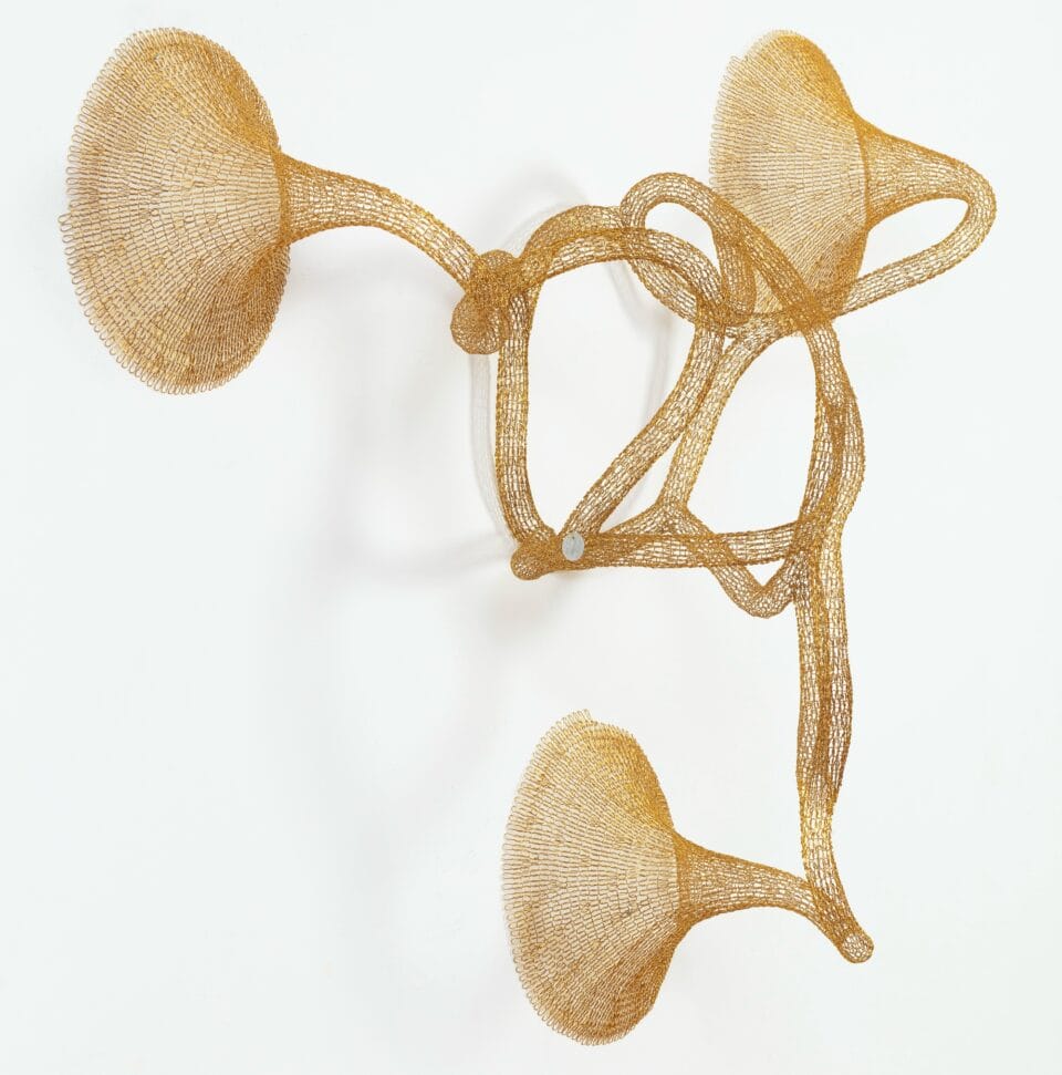 a gold abstract woven metal sculpture with tubes and circular forms on a wall