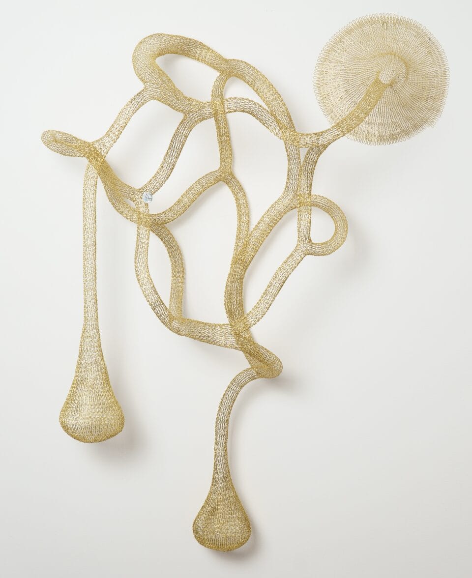 a gold abstract woven metal sculpture with tubes and circular forms on a wall
