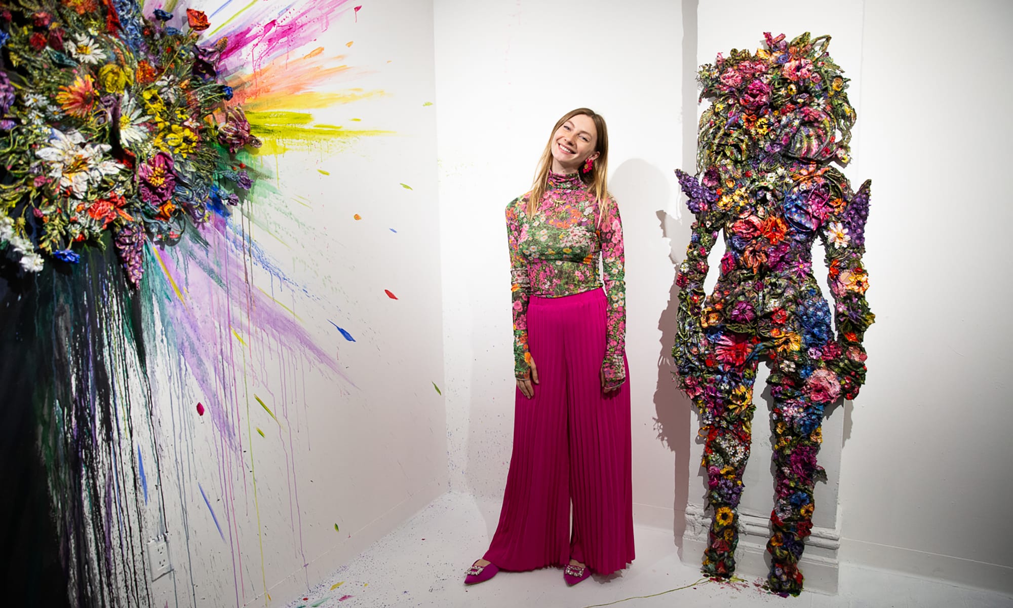 a white woman in a floral top and pink wide leg pants poses with a floral figure and a bursting floral wall work