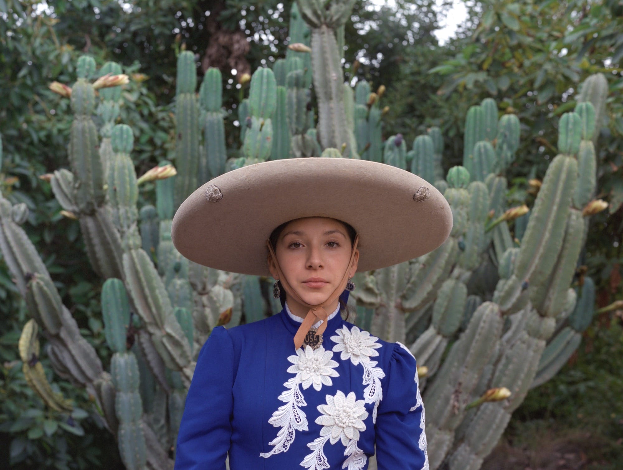 a woman stands in front of a cactus, wearing a blue-and-white top and a wide-brimmed hat