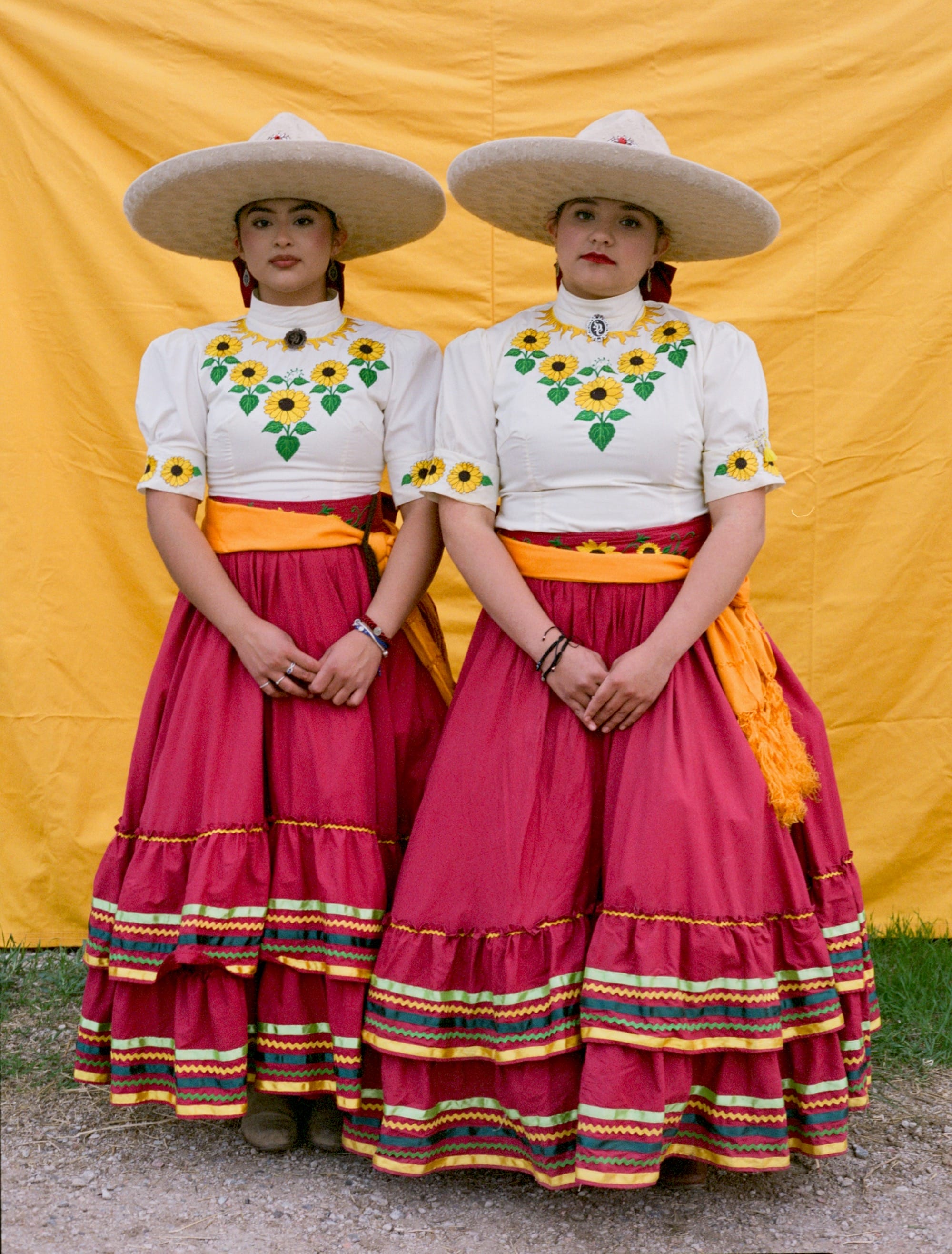 two women stand side-by-side in front of a yellow backdrop, wearing magenta and white skirts and wide-brimmed hats