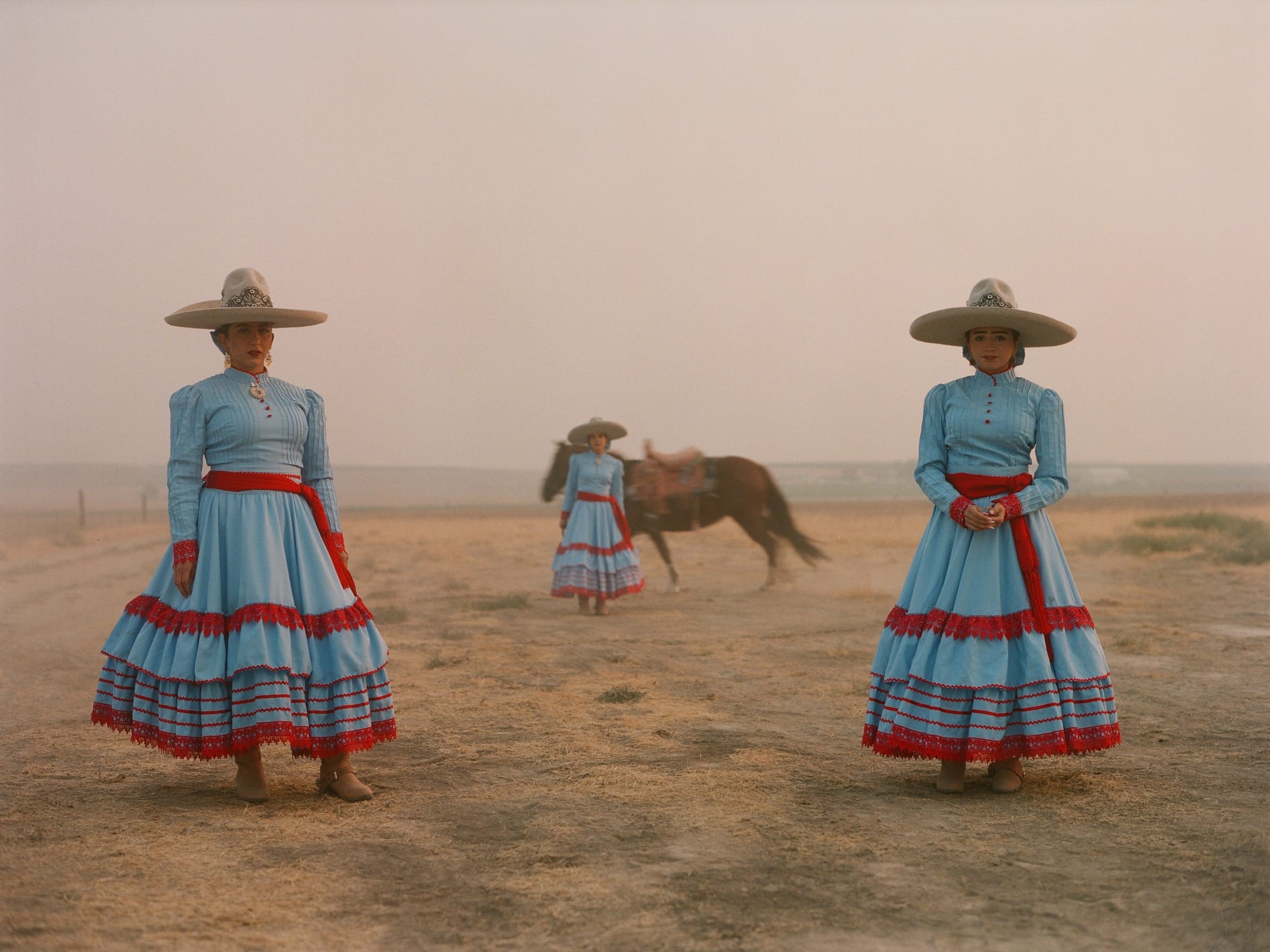 three women in colorful dresses and wide-brimmed hats stand in the desert, with a horse running in the background