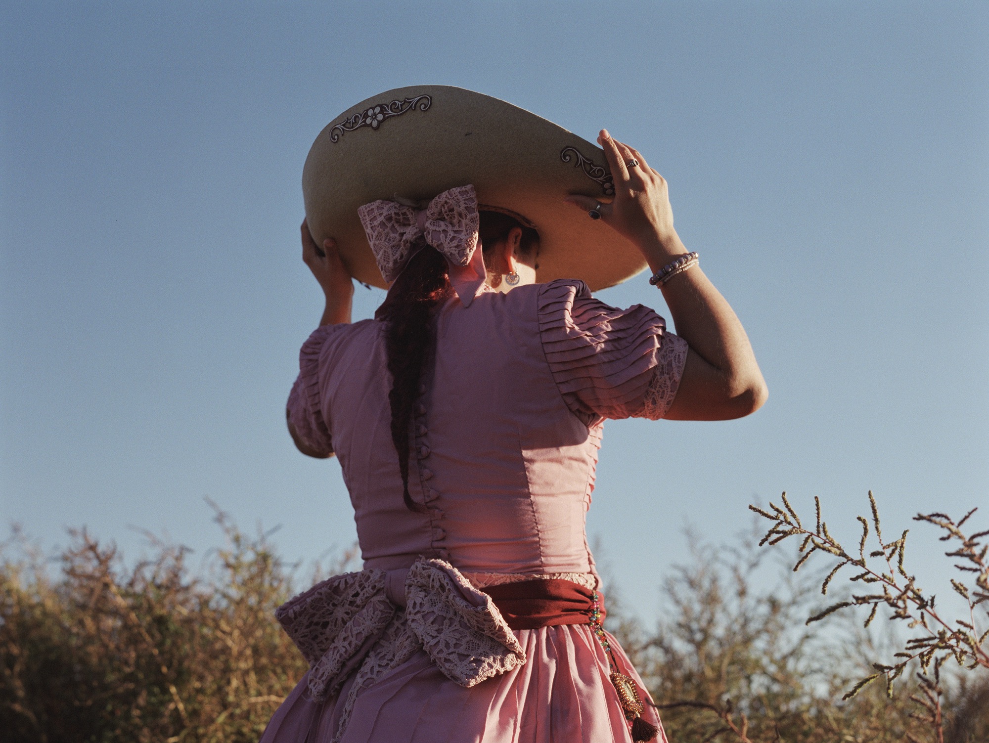 a young woman is seen from the back in a pink dress, adjusting a wide-brimmed hat