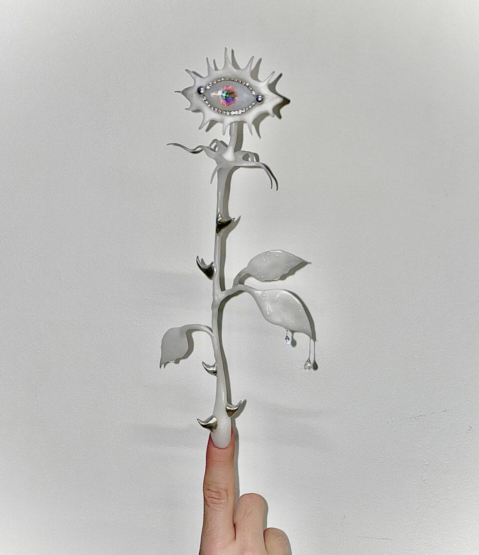 a pointer finger shows a very long all-white flower design extending upward. the stem is thorny with leaves and the petals are spiky and surround a rhinestoned eye
