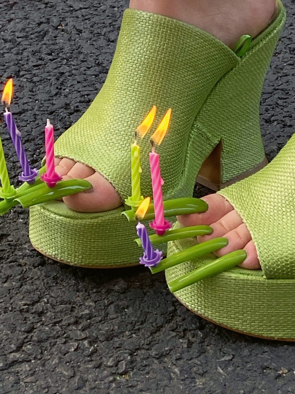 a pair of feet in chunky green heels sporting long green acrylic nails with lit birthday candles on the ends