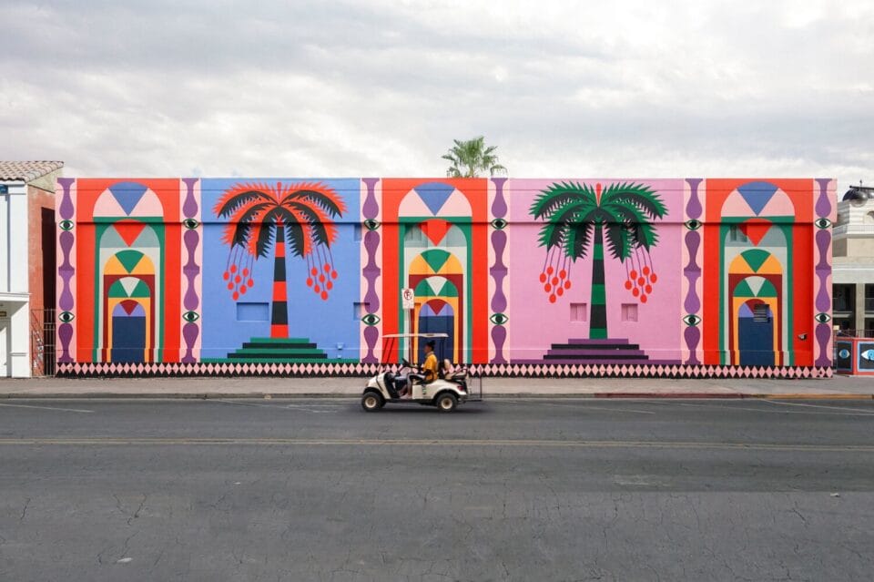 a long, wide building with colorful murals of palm trees and geometric patterns, photographed with a person riding a scooter in front of it