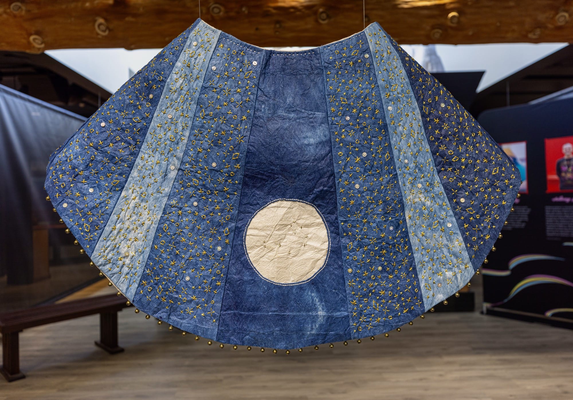 a barkcloth tapestry installation with indigo-dyed panels in different shades, sewn together to create a ray-like effect, with a metallic circle in the bottom center
