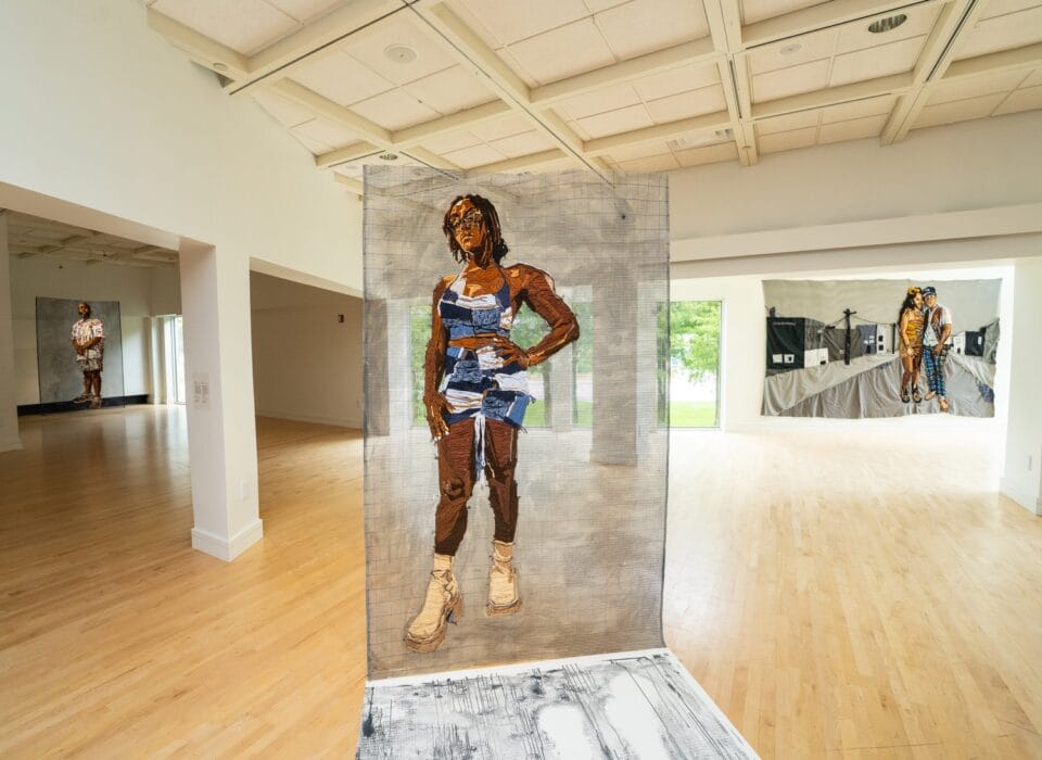 a large-scale embroidered portrait of a vibrantly-dressed Black woman against a gridded, semi-transparent mesh background, installed in a gallery space