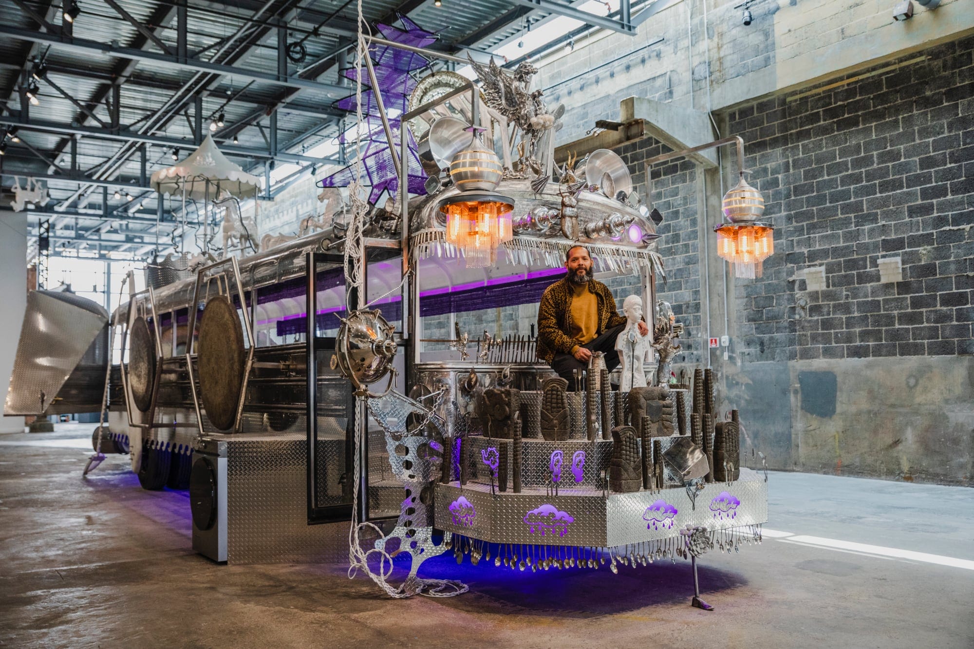 the artist sits on a chrome school bus sculpture