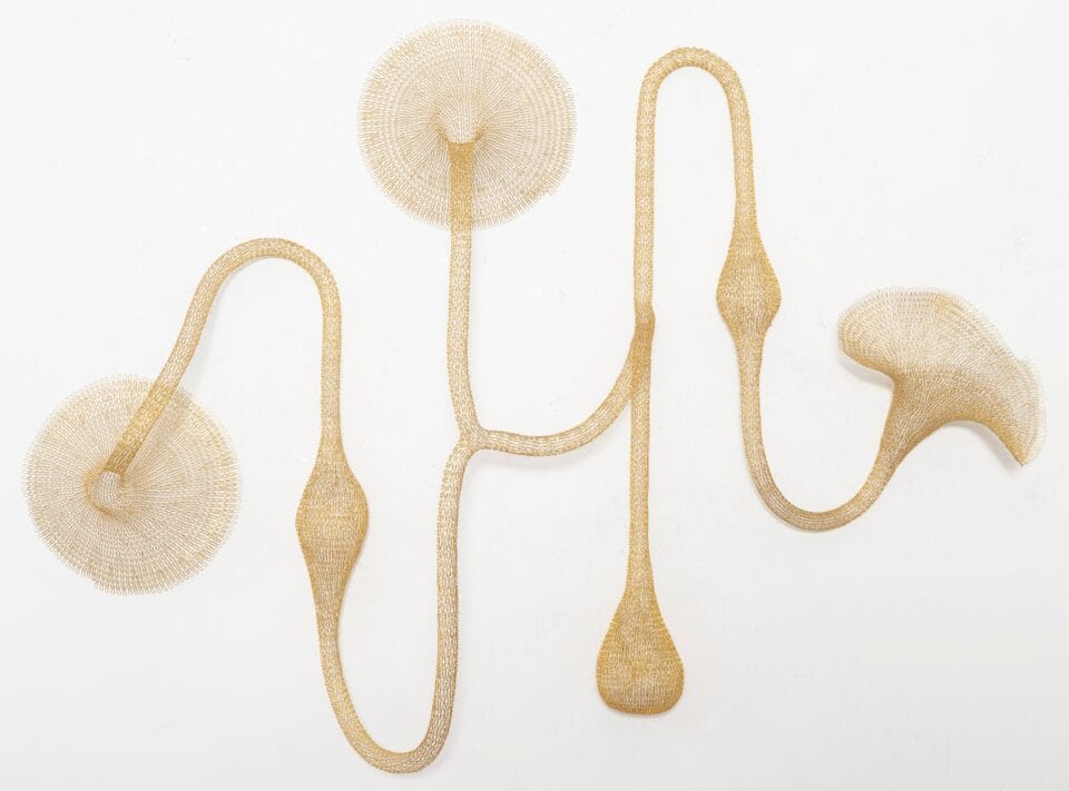 a gold abstract woven metal sculpture with tubes and circular forms on a wall