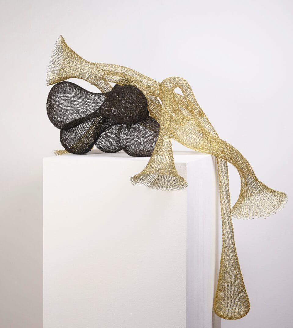 a gold and black abstract woven metal sculpture with tubes and circular forms spilling over a pedestal