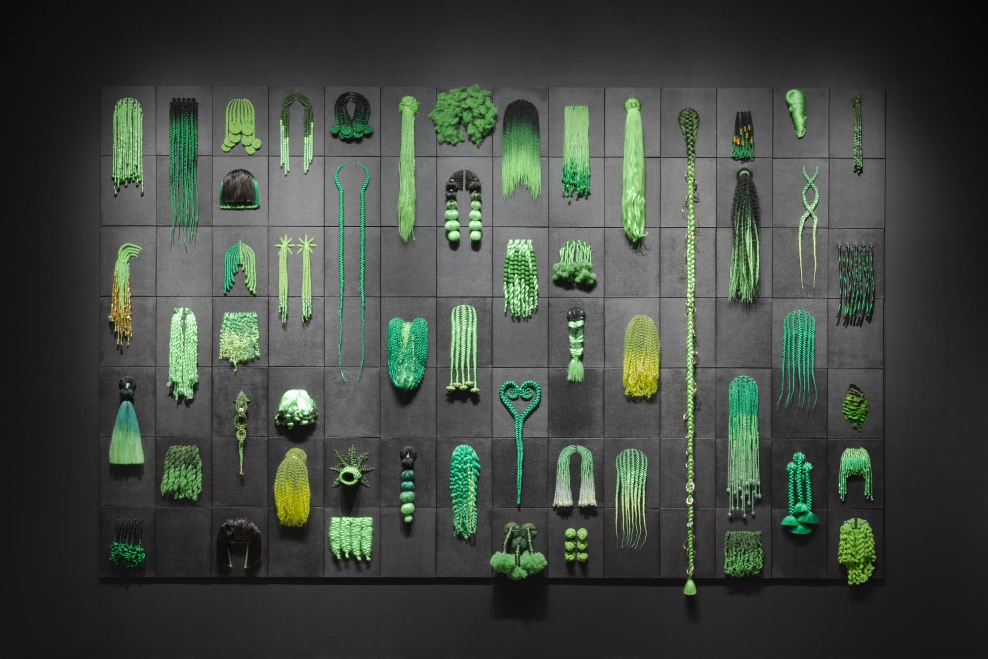 an art installation of numerous green wigs in a variety of braided styles on a black wall