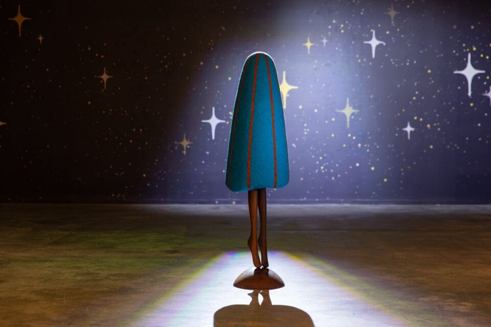 an abstract, colorful, soft sculpture in a darkened gallery installation, with legs as if the form is dancing