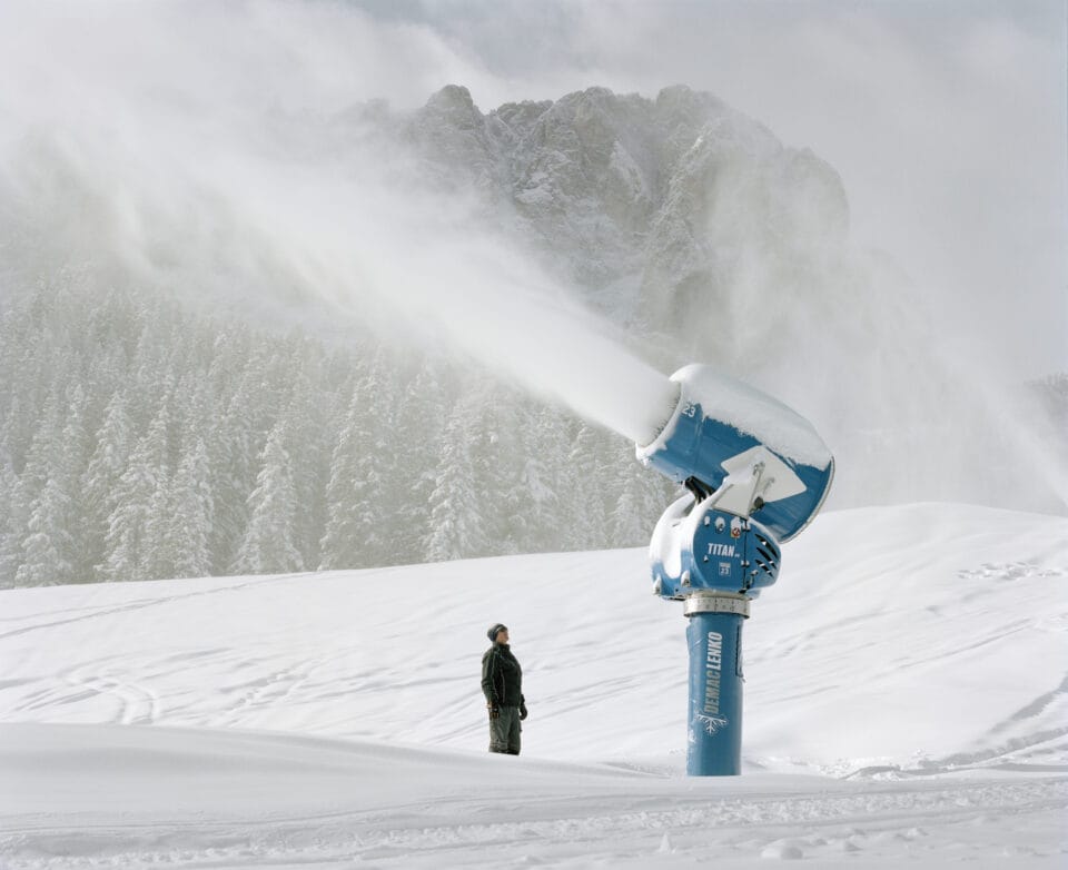 a man stands beside a giant snow cannon throwing artificial snow in a snowy mountain scene