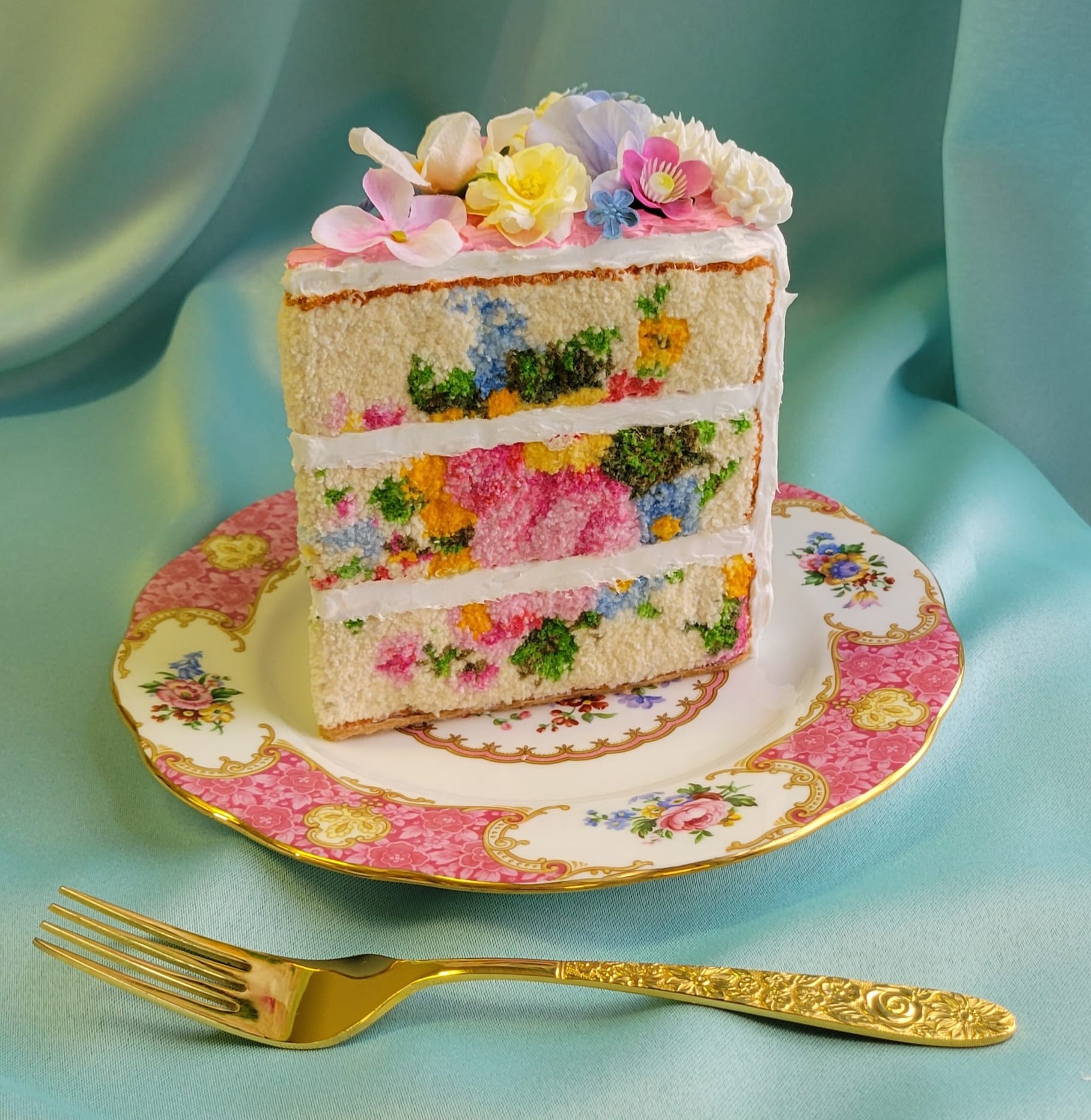 a small sculpture of a slice of layer cake on an ornate pink plate, next to a gold fork, with realistic frosting details and embroidery on that looks like the texture of fluffy sponge