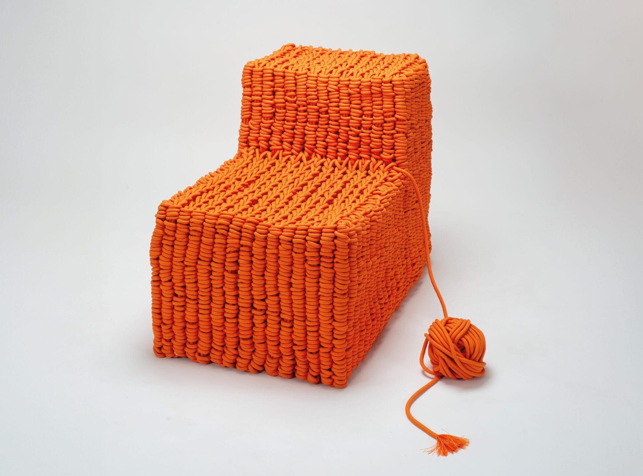 a geometric, armless chair made of orange nylon rope, with a ball of the rope connected to it and sitting by its side on the floor
