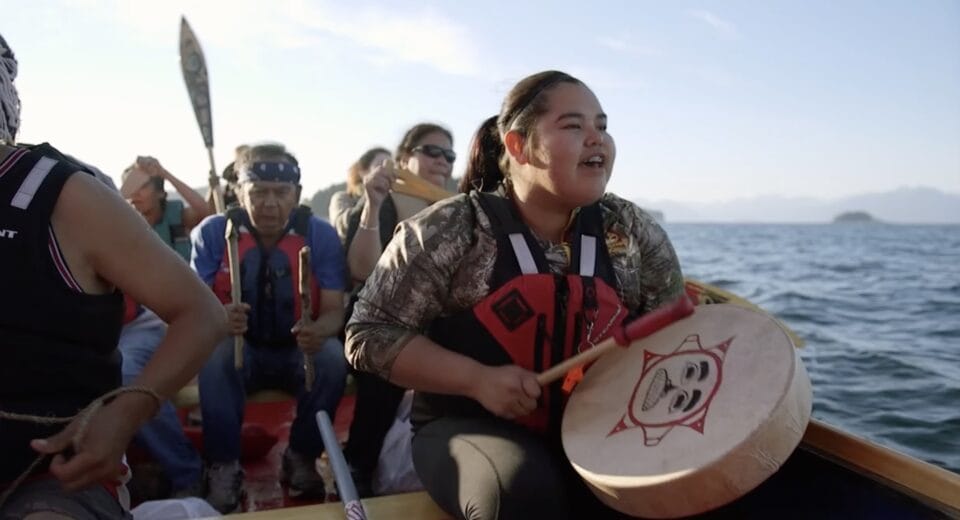 a still from a short film about a Huna Tlingit ancestral tribal house being constructed, showing a woman in a boat with others, banging a ceremonial drum and singing