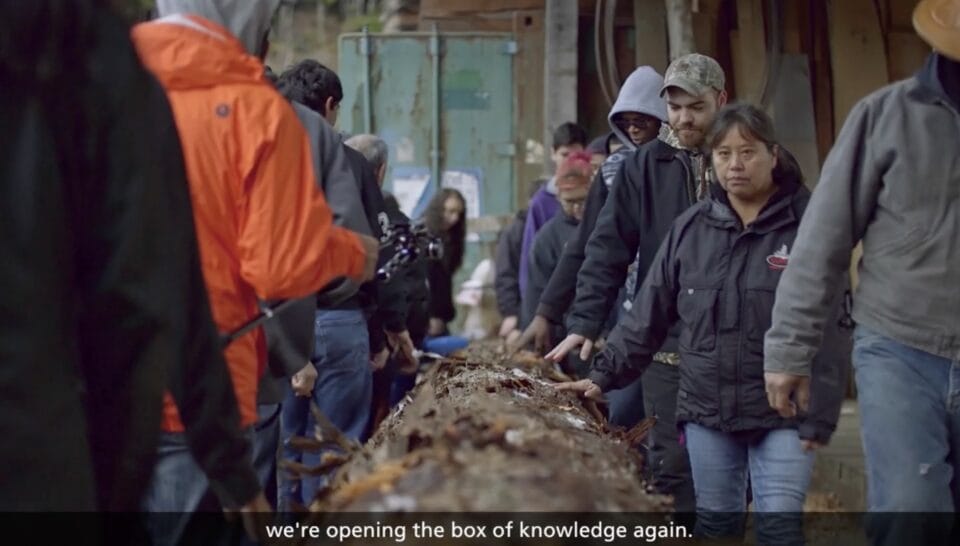 a still from a short film about a Huna Tlingit ancestral tribal house being constructed, showing local residents touching a large tree that has been felled for a totem pole